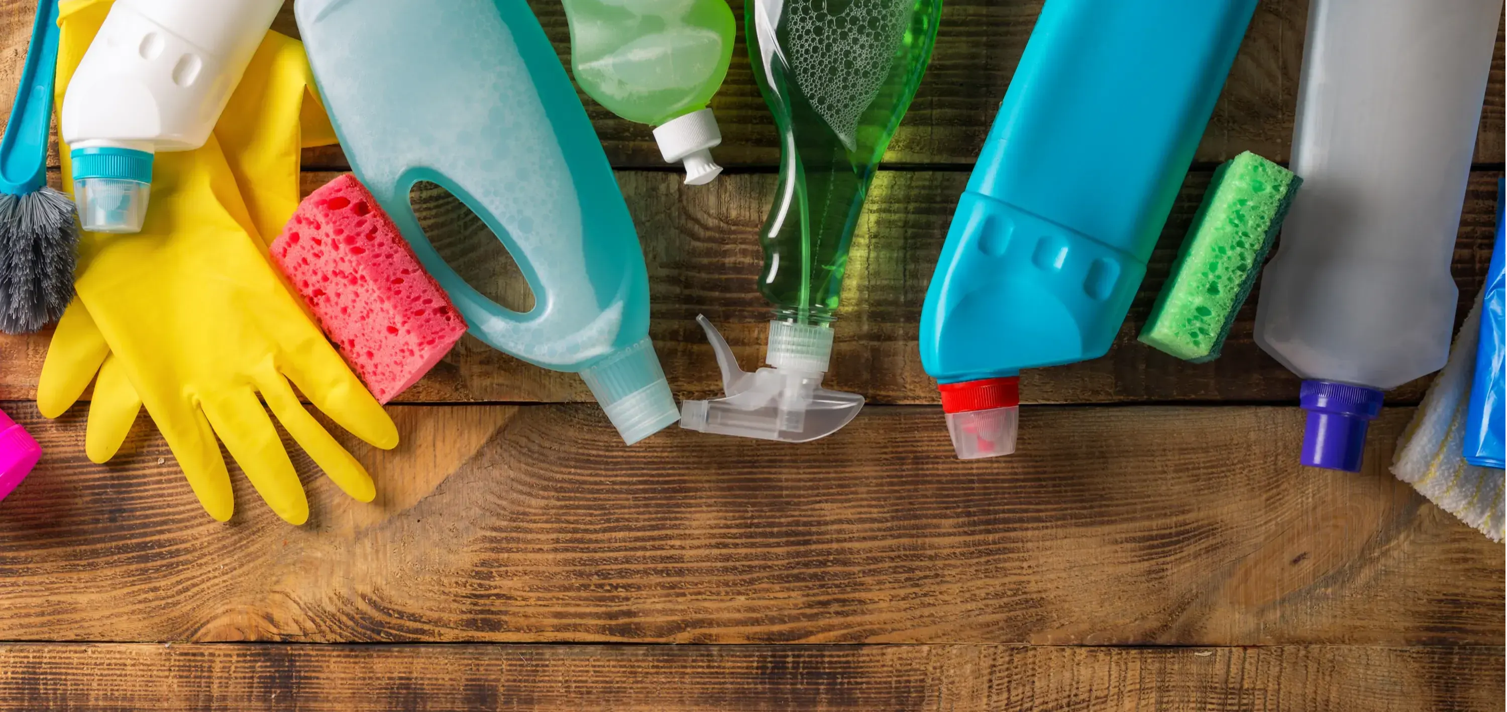 A variety of cleaning supplies on a wooden table. Displayed are spray bottles, sponges, cloths, and a bucket.